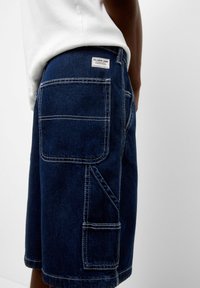 Pantalones cortos de mezclilla azul oscuro con costuras en contraste, que cuentan con múltiples bolsillos, una etiqueta de marca en la cinturilla y un corte recto.