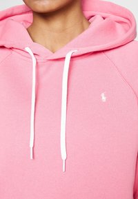 Rosa bomullshoodie med vita dragsko, med en liten vit logotyp på vänster bröst och ribbade detaljer vid kanterna.