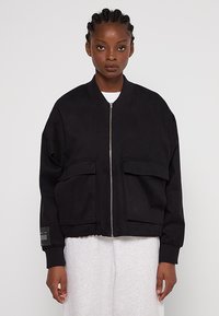 Czarna kurtka bombowiec z grubego materiału, z frontowym zamkiem błyskawicznym, dwoma dużymi naszywanymi kieszeniami i ściągaczami na mankietach. Na rękawie znajduje się metka.