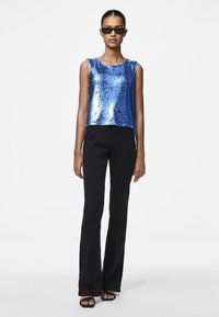 Top à sequins bleu avec un col rond, assorti à un pantalon évasé noir et des sandales plates. Le modèle porte des lunettes de soleil noires.