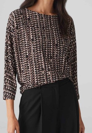 Mujer con blusa marrón con patrones verticales de puntos en blanco y negro, y pantalones negros de talle alto.