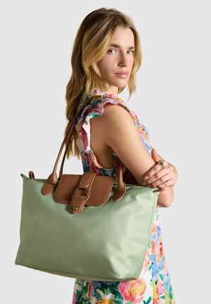 Blonde woman in floral dress carries large light green tote bag with brown leather straps and buckle over her shoulder, looking sideways.