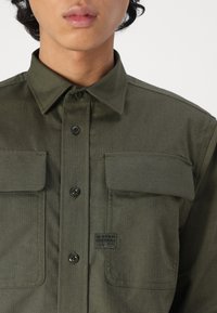 Olijfgroene shirt met een button-down kraag, twee borstzakken en een contrasterend label. Gemaakt van geweven stof met een gestructureerde afwerking.