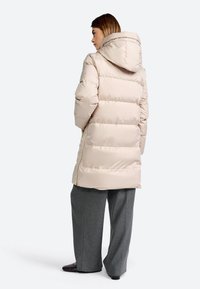 Beige puffer jas met capuchon, gewatteerd ontwerp en zijritsen. Gekoeld met grijze wijde broek en donkere schoenen.