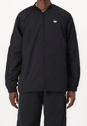 Mężczyzna ubrany w czarną rozpinaną kurtkę z elastycznymi mankietami i małym białym logo Adidas na lewej piersi, w połączeniu z czarnymi spodniami.