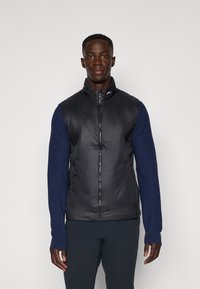 Čierna vypchávkovaná vesta s vysokým golierom a zipsom vpredu, spárovaná s tmavomodrými dlhými rukávmi. Hladká textúra s kontrastnými materiálmi a jemným logom.