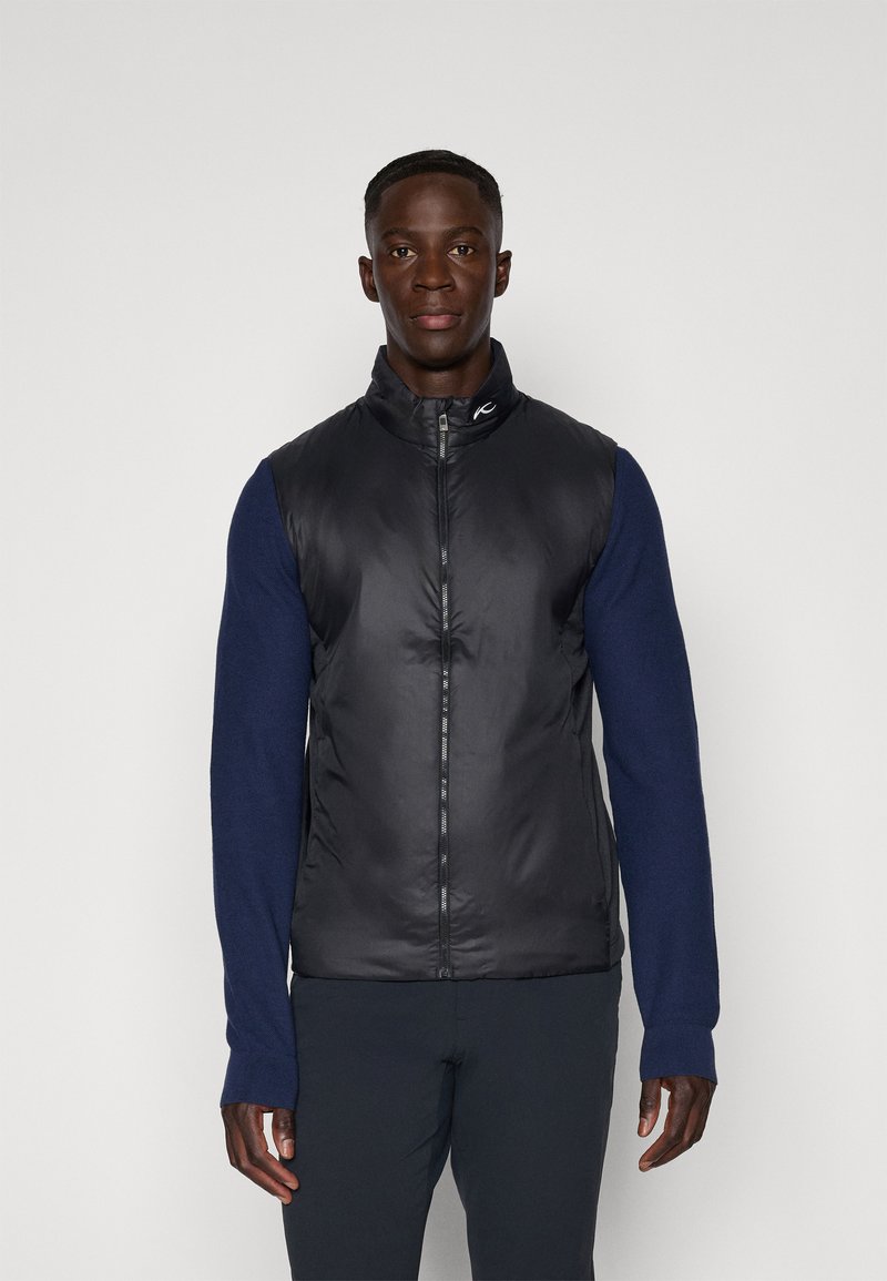 Čierna vypchávkovaná vesta s vysokým golierom a zipsom vpredu, spárovaná s tmavomodrými dlhými rukávmi. Hladká textúra s kontrastnými materiálmi a jemným logom.