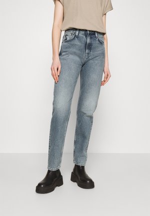 Persoon draagt high-waist grijs gewassen spijkerbroek, beige korte mouw top en zwarte chunky platform enkellaarsjes tegen een effen achtergrond.