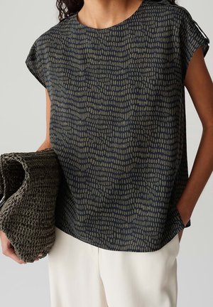 Vrouw die een zwart-olijfgroen kortarmig top met patroon draagt, een grote donkere geweven clutch vasthoudt, met zichtbare witte broek.