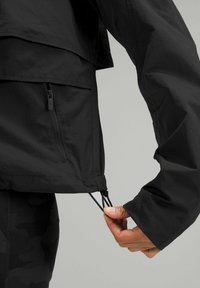 Main ajustant le cordon de l'ourlet de la veste noire près de la poche zippée latérale, portant des leggings noirs sur un fond neutre.