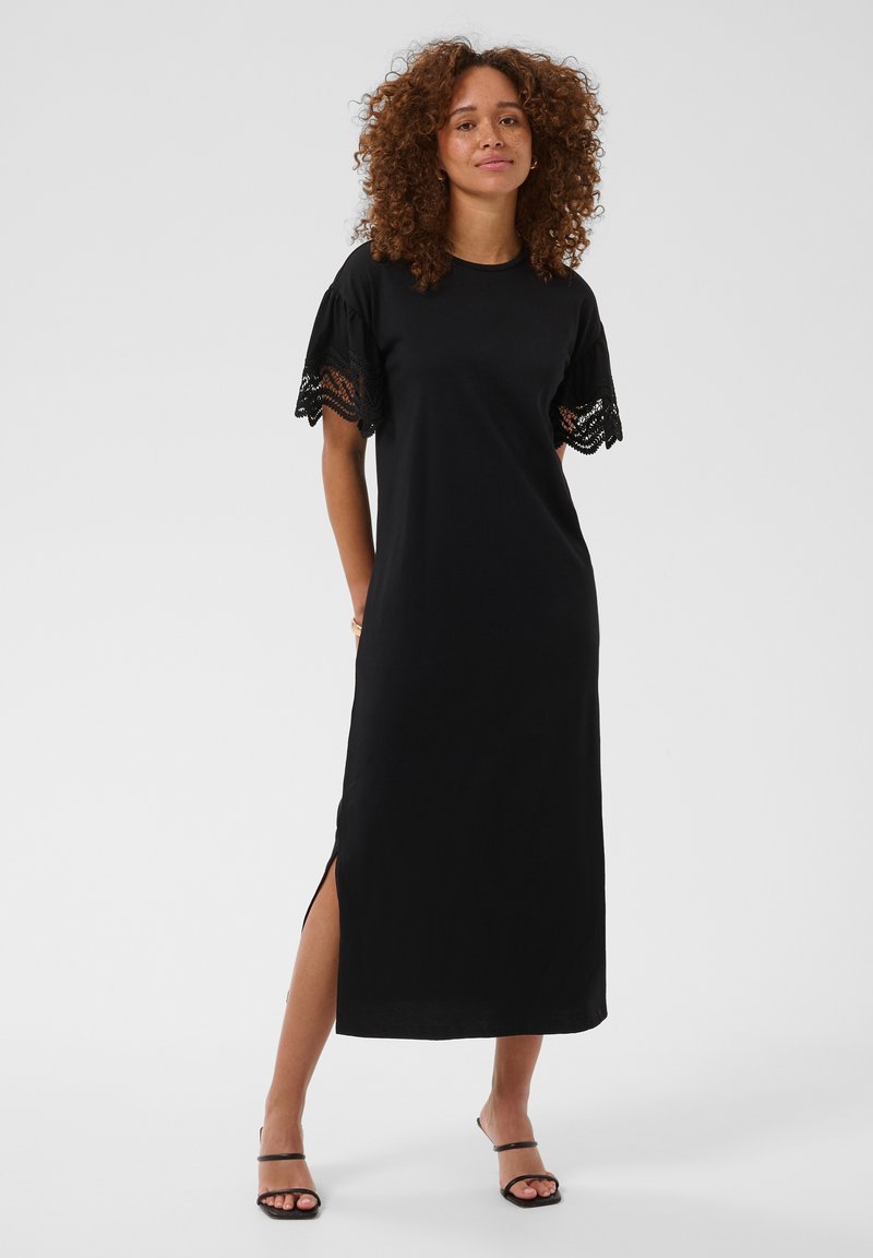 Femme debout dans une robe midi noire à manches courtes en dentelle avec une fente latérale, portant des talons noirs à brides sur un fond uni.