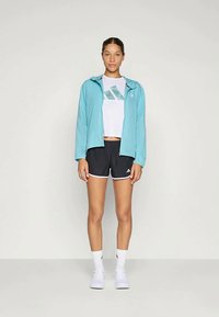 Hellblaues Zip-Up-Jackett mit Kapuze, weißes T-Shirt mit kurzen Ärmeln und grünem Grafikmotiv, schwarze Shorts mit weißem Saum und weiße Sportsocken.