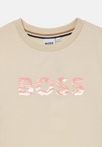 Bež t-shirt iz bombaža z okroglo izrezom. Ponuja sijoč roza logotip "BOSS" z teksturiranim vzorcem. Diskretni notranji barvni poudarki.