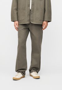 Άνδρας που φοράει φαρδιά ελιάς πράσινης απόχρωσης παντελόνια, αντίστοιχο σακάκι με μεγάλες μπροστινές τσέπες και εκρού αθλητικά παπούτσια με σόλες από καουτσούκ.