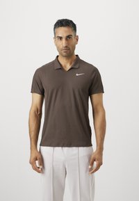 Muž stojí čelom dopredu v hnedom Nike polo tričku s krátkym rukávom a svetlých béžových nohaviciach na jednolatom pozadí.