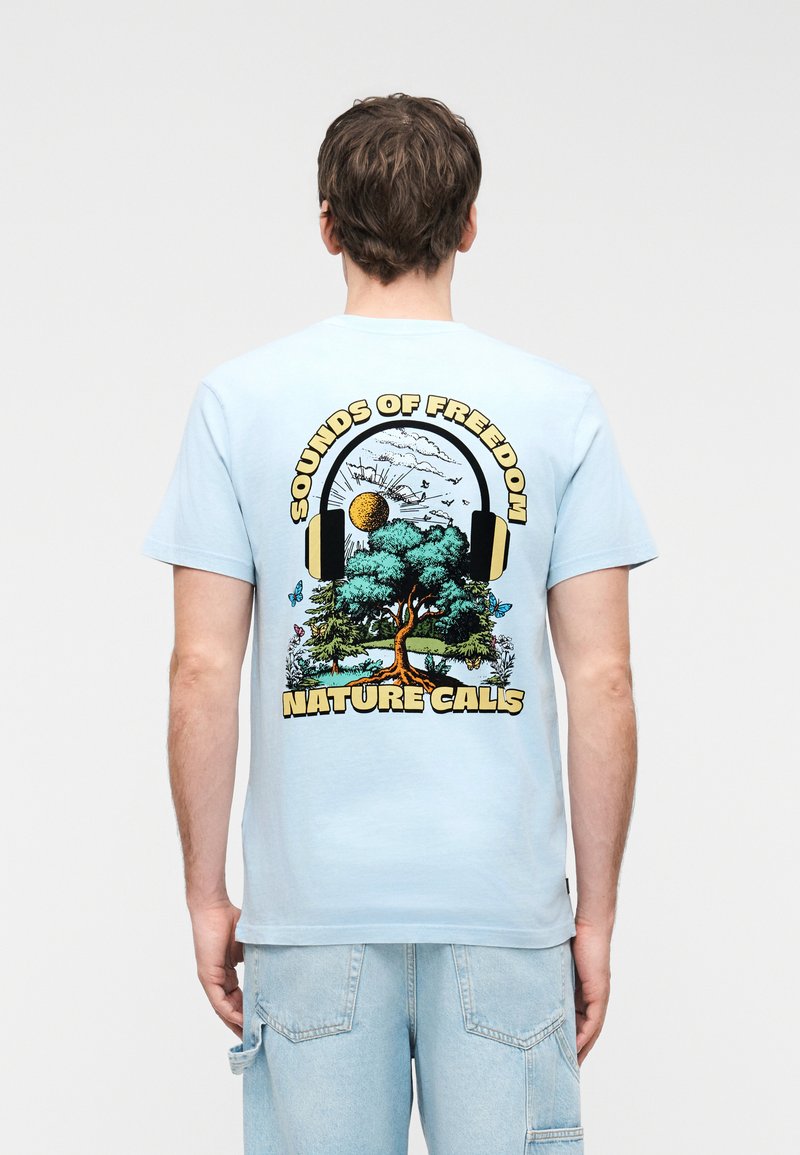 Άνδρας που φοράει ανοιχτό μπλε μπλουζάκι με γραφικό σχεδιασμό δέντρου, ήλιου, πουλιών και ακουστικών, με το κείμενο «Sounds of Freedom Nature Calls» στην πλάτη.