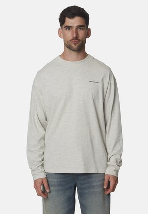 Man met lichtgrijs shirt met lange mouwen met kleine tekst op de borst en blauwe spijkerbroek, staand tegen een effen lichte achtergrond.