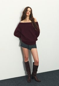 Kobieta o długich, falowanych włosach, ubrana w bordowy sweter z odsłoniętymi ramionami, ciemne szorty i brązowe, skórzane kozaki do kolan, stojąca na tle białej ściany.