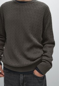 Maglione marrone a trecce con scollatura rotonda, con polsini e orlo a costine. Indossato sopra uno strato blu scuro, abbinato a jeans grigi.