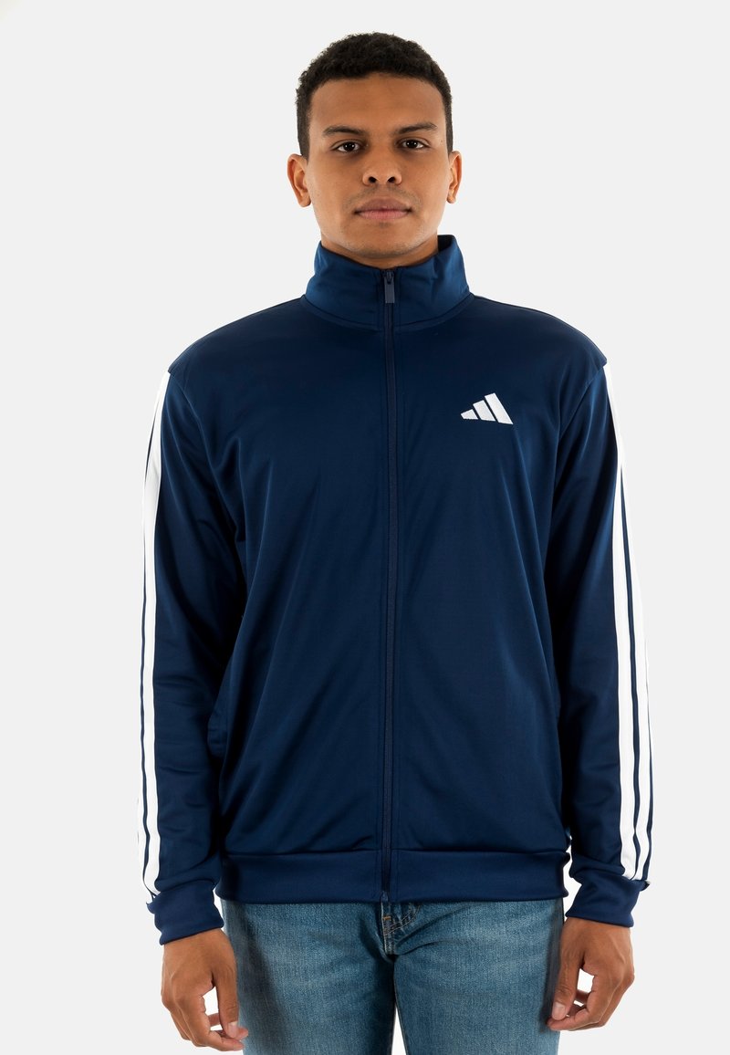 Marineblauwe sportjack met rits en hoge kraag, wit Adidas-logo op de borst en witte strepen langs de mouwen. Glad, rekbaar materiaal.