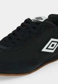 Baskets Umbro noires avec lacets noirs, logo blanc sur le côté et la languette, et semelle en caoutchouc marron.