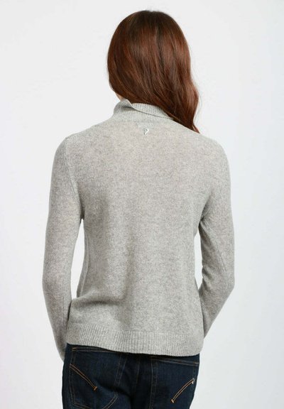 Donna con lunghi capelli castani, che indossa un maglione grigio chiaro a maniche lunghe e jeans blu scuro, vista di spalle su uno sfondo neutro.