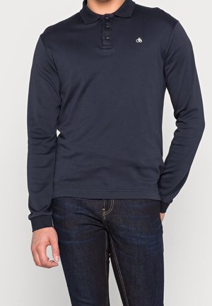 Mann trägt ein marineblaues, langärmliges Polohemd mit drei Knöpfen und dunkelblaue Jeans, steht mit einer Hand in der Tasche.