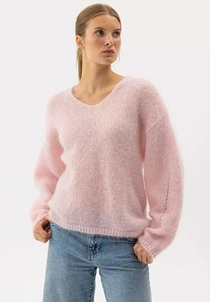 Giovane donna che indossa un maglione rosa chiaro peloso con scollo a V e jeans blu, in piedi con le mani nelle tasche, guardando di lato.