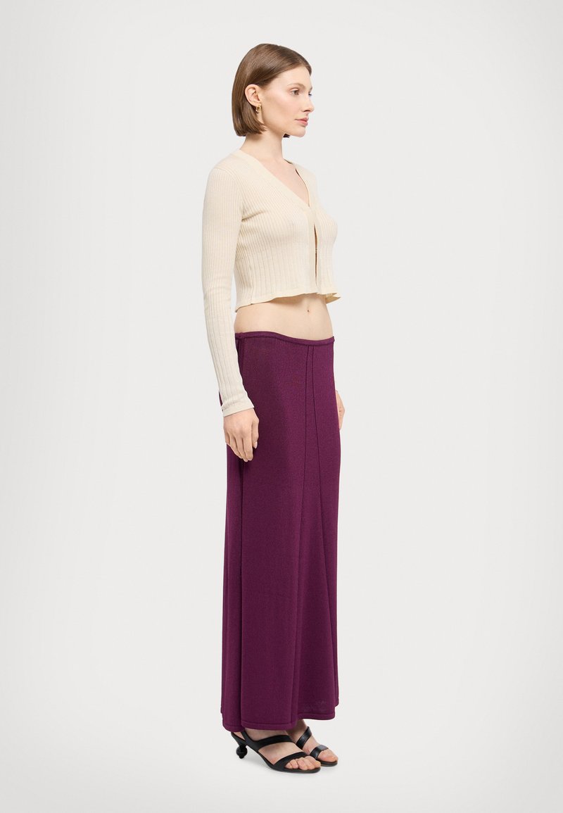 Cardigan scurt ribatat bej cu mâneci lungi, asortat cu o fustă lungă, fluidă de culoare mov. Modelul poartă sandale negre.