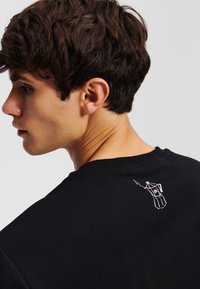 Juodas medvilninis marškinėlis, kuriame nugaros pusėje pavaizduotas mažas baltas kavos virimo aparato kontūras. Trumpomis rankovėmis su klasikiniu apvaliu kaklu.