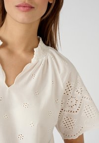 Blusa bianca di cotone con scollatura arricciata, maniche corte e delicati ricami ad occhiello con motivi floreali.