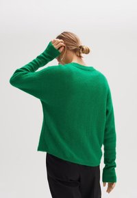 Zielony sweter wykonany z miękkiego materiału, o luźnym kroju, z długimi rękawami i ściągaczami przy mankietach. Widok z tylu pokazuje zaokrąglony dół.