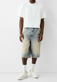 Camiseta blanca corta, pantalones cortos de mezclilla deslavados en azul claro con dobladillo deshilachado y zapatillas deportivas blancas con suelas gruesas y cordones.