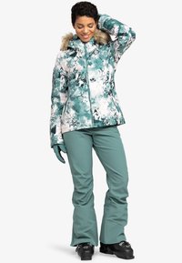 Winterjacke met een teal en witte bloemmotief en een nepbont capuchon, gecombineerd met bijpassende teal broeken en zwarte skISchoenen.