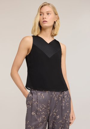 Donna bionda che indossa un top nero senza maniche con scollo a V strutturato e pantaloni grigi con motivo floreale, in piedi con una mano in tasca.