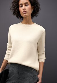 Giovane donna con capelli ricci che indossa un maglione crema e pantaloni a scacchi bianco e nero su uno sfondo grigio scuro.