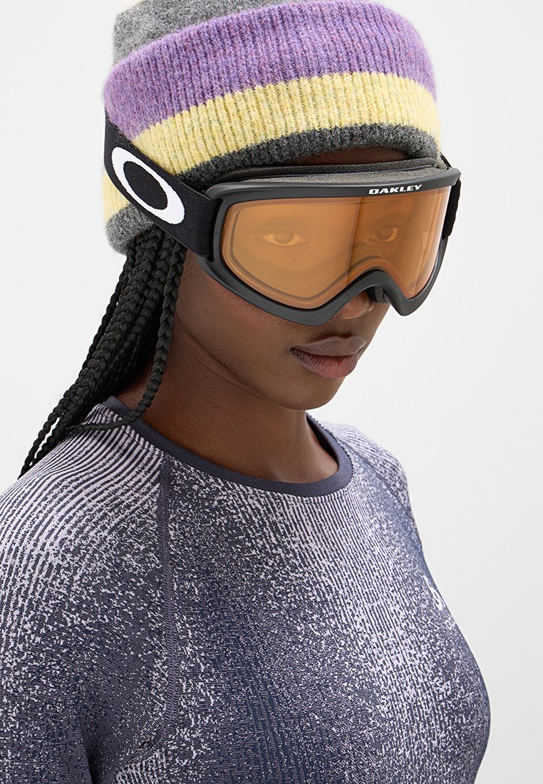 Zwarte skibril met oranje lenzen, voorzien van een dikke schuimvoering en een verstelbare band. Grijs en paars gestreepte gebreide beanie gedragen bovenop.