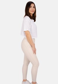 Weißes gekürztes T-Shirt kombiniert mit beigen Leggings, die eine glatte Textur und eine schlanke Passform haben. Das Model steht seitlich und trägt weiße Sneakers.