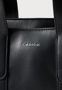 Juodas odinis rankinis krepšys su lygiu paviršiumi, kuriame yra sidabrinė "Calvin Klein" logotipas. Apima struktūruotas rankenas ir minimalistinį dizainą.