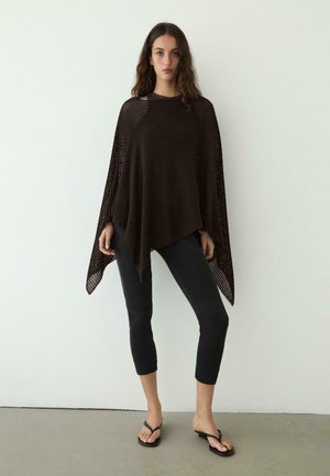 Femme portant un poncho en maille ample marron foncé, un pantalon noir court et des sandales tongs noires, debout contre un mur blanc uni.