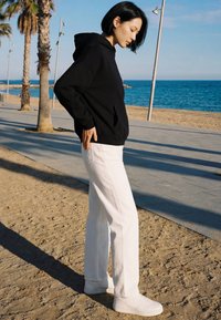 Jeune femme en sweat à capuche noir et pantalon blanc sur une promenade de plage de sable près des palmiers et de l'océan sous un ciel clair.