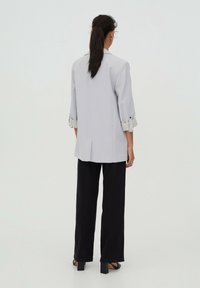 Femme aux cheveux bruns attachés en queue de cheval, portant un blazer gris clair aux manches retroussées, un pantalon noir à jambes larges et des sandales à talons noires, vue de dos.