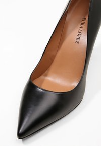 Černé koženkové lodičky na vysokém podpatku s špičatou špičkou, hladkým povrchem a nahým interiérem. Výrazná značka: "PURA LÓPEZ" embosovaná uvnitř.