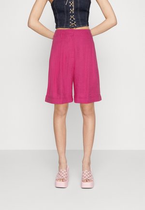 Mujer con shorts Bermuda de talle alto en un brillante color rosa combinados con sandalias de plataforma a cuadros rosas y una blusa sin mangas negra con cordones.