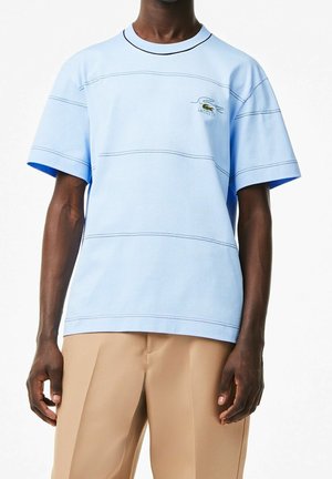 Lyseblå kortærmet t-shirt med kontrastfyldt mørk syning og broderet grønt Lacoste-logo. Bruges med beige bukser. Blød tekstur.