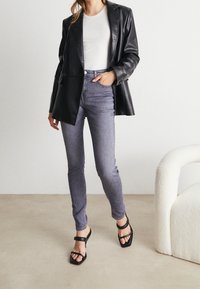 Blazer de cuero negro sobre una camisa blanca ajustada, combinado con jeans skinny gris de cintura alta y sandalias negras con tiras. Texturas suaves y diseño ajustado.