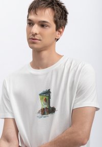 Witte katoenen T-shirt met een afbeelding van een groene dumpster met verschillende voorwerpen eromheen, waaronder een cartoonachtig gezicht en gevarieerde texturen.