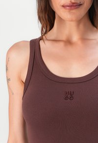 Débardeur marron à côtes avec un col rond et un petit logo brodé "HU GO" en bordeaux au centre. Vue en gros plan.
