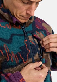 Fleece bunda s vícebarevným abstraktním vzorem v petrolejové, hnědé a fialové barvě. Má vysoký límec a kapsu na zip na hrudi.