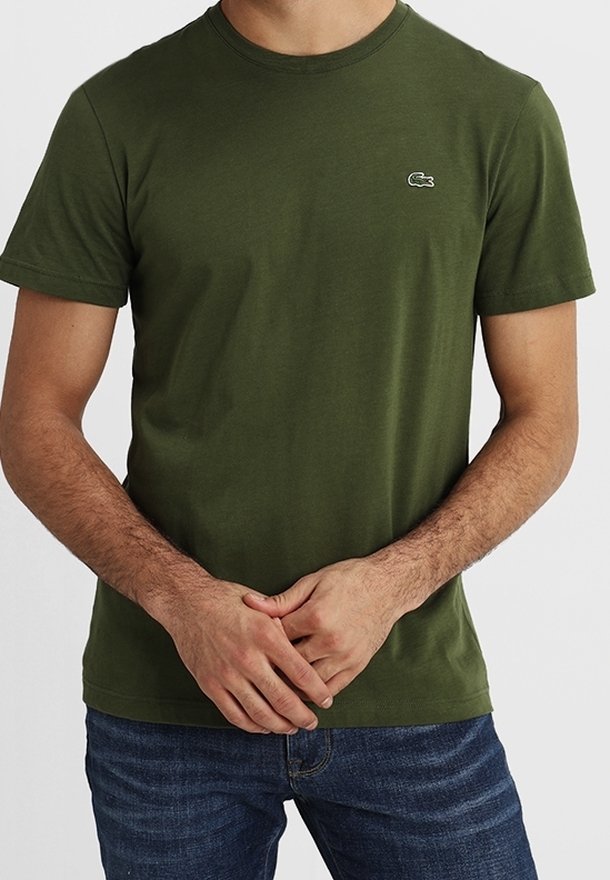 Homme portant un t-shirt uni vert olive à manches courtes avec un petit logo crocodile sur la poitrine et un jean bleu, mains jointes devant.